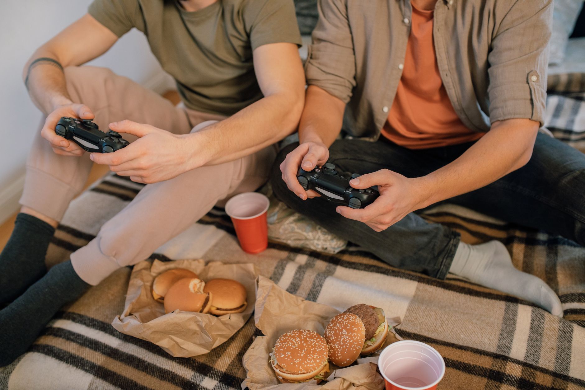 Deux personnes assises sur une couverture à carreaux tiennent des manettes noires, avec des hamburgers sur du papier kraft et des gobelets rouges.